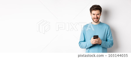 Smiling modern man using smartphone and looking pleased at camera. Guy in sweatshirt with mobile phone on white background 103144035