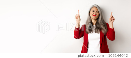Skeptical and disappointed asian businesswoman pointing fingers up, grimacing and showing bad promo offer, standing over white background 103144042