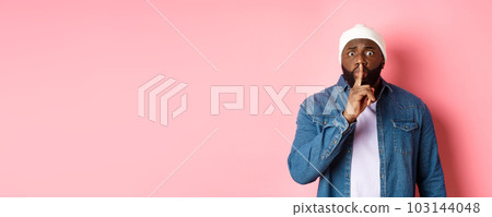 Worried Black man asking to keep quiet, sharing secret and hushing at you, holding finger pressed to lips and staring nervously at camera, pink background Worried Black man asking to keep quiet, sharing secret and hushing at you, holding finger pressed to lips and staring nervously at camera, pink background 103144048