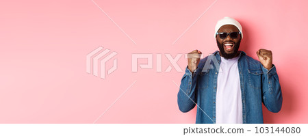 Satisfied african-american man in sunglasses saying yes, triumphing and making fist pumps in rejoice, celebrating achievement, standing like winner over pink background 103144080
