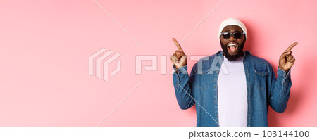 Amazed cool Black guy showing two variants, pointing fingers sideways and staring at camera impressed, wearing sunglasses, standing over pink background Amazed cool Black guy showing two variants, pointing fingers sideways and staring at camera impressed, wearing sunglasses, standing over pink background 103144100