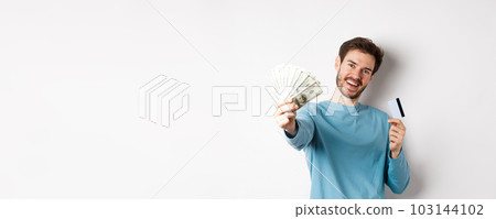 Handsome young man in blue shirt giving you money, holding plastic credit card, standing over white background Handsome young man in blue shirt giving you money, holding plastic credit card, standing over white background 103144102