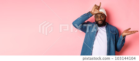 Sad african-american man showing loser sign on forehead and staring at camera, standing over pink background Sad african-american man showing loser sign on forehead and staring at camera, standing over pink background 103144104