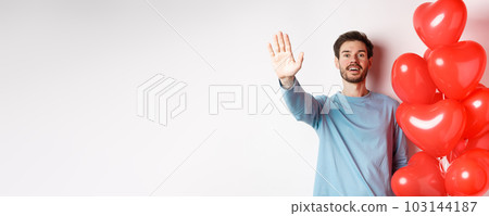 Handsome young caucasian man standing with romantic heart balloon and waving hand at lover, waiting for his date on Valentines day with cute surprise, standing over white background 103144187