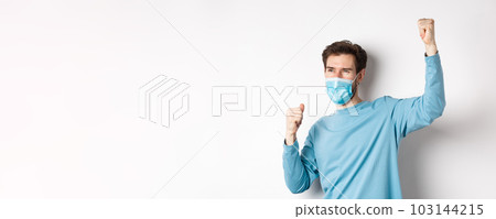 Covid-19, pandemic and social distancing concept. Happy man in medical mask watching sports game and cheering, raising hands up to celebrate, standing over white background Covid-19, pandemic and social distancing concept. Happy man in medical mask watching sports game and cheering, raising hands up to celebrate, standing over white background 103144215