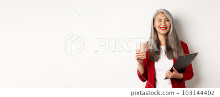 Successful asian businesswoman drinking coffee and winking cheerful at camera, standing with clipboard, wearing red blazer, white background Successful asian businesswoman drinking coffee and winking cheerful at camera, standing with clipboard, wearing red blazer, white background 103144402