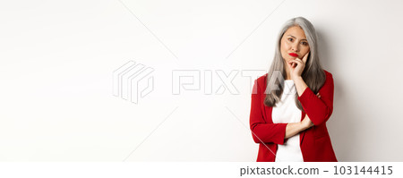 Annoyed and bothered asian businesswoman in red blazer, pouting and looking irritated at camera, standing over white background Annoyed and bothered asian businesswoman in red blazer, pouting and looking irritated at camera, standing over white background 103144415