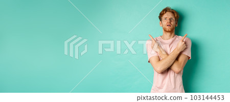 Confused young man in t-shirt looking up, pointing fingers sideways and feeling puzzled, cant decide, standing over turquoise background Confused young man in t-shirt looking up, pointing fingers sideways and feeling puzzled, cant decide, standing over turquoise background 103144453