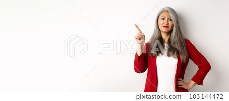 Skeptical asian businesswoman in red blazer grimacing, pointing upper left corner and looking dissatisfied, disapprove something, standing over white background 103144472
