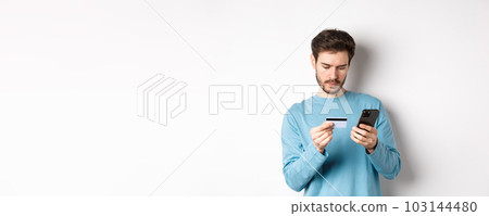 E-commerce and shopping concept. Young man making online payment, holding plastic credit card and smartphone, standing on white background E-commerce and shopping concept. Young man making online payment, holding plastic credit card and smartphone, standing on white background 103144480