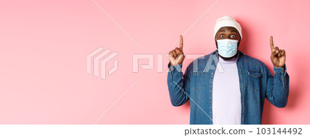 Coronavirus, lifestyle and global pandemic concept. Excited african-american guy in medical mask showing announcement, pointing fingers up, standing over pink background 103144492
