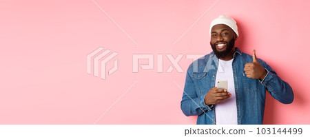 Technology and online shopping concept. Happy african-american guy showing thumb-up in approval while using mobile phone, standing satisfied over pink background 103144499