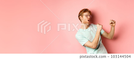 Cheerful redhead guy listening music and dancing, triumphing or celebrating success, standing over pink background 103144504