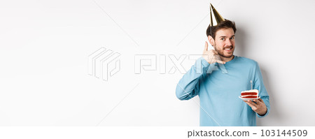 Celebration and holidays concept. Cheerful smiling man in party hat, celebrating birthday with bday cake, showing call me phone gesture near ear, white background 103144509