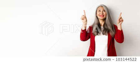Happy asian lady in red blazer and makeup, looking and pointing fingers up, showing special offer, standing over white background 103144522