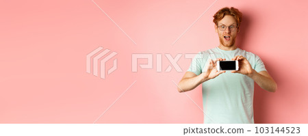 Amazed redhead man gasping and staring with awe phone, showing blank smartphone screen horizontally, standing over pink background 103144523