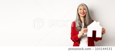 Asian female real estate agent showing paper house cutout, broker smiling friendly and selling property, standing over white background 103144531