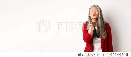 Elegant asian woman with red blazer and lips, blowing air kiss at camera, concept of valentines day and romance, white background 103144546
