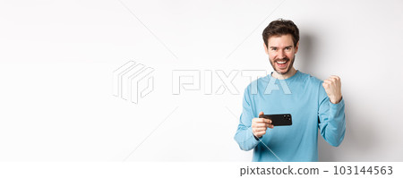 Lucky man winning in mobile video game, holding smartphone and saying yes, triumphing with fist pump, standing over white background 103144563