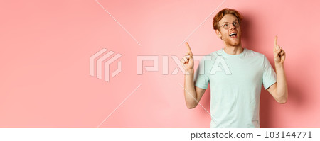 Amazed redhead guy in glasses checking out special deal, looking and pointing fingers up with amazed face, standing over pink background Amazed redhead guy in glasses checking out special deal, looking and pointing fingers up with amazed face, standing over pink background 103144771