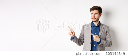 Handsome businessman in suit pointing, looking left at logo with serious face, standing against white background 103144849