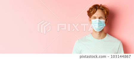 Covid-19 and pandemic concept. Close up of redhead man in medical mask looking at camera, standing over pink background 103144867