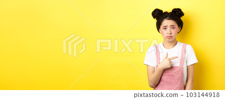 Sad and disappointed asian girl frowning, feel unfair, pointing finger right at bad thing, complaining on something upsetting, yellow background Sad and disappointed asian girl frowning, feel unfair, pointing finger right at bad thing, complaining on something upsetting, yellow background 103144918