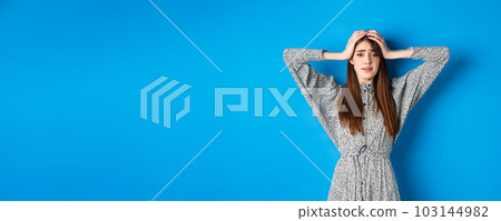 Frustrated adult woman with natural hair holding hands on head and panicking, looking alarmed and scared, standing in dress against blue background 103144982