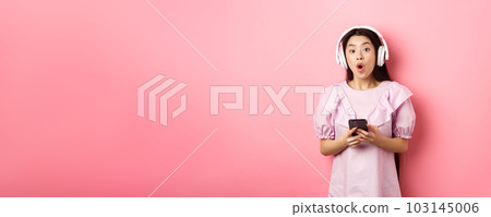Excited asian woman in wireless headphones say wow, holding mobile phone and looking amused at camera, standing against pink background Excited asian woman in wireless headphones say wow, holding mobile phone and looking amused at camera, standing against pink background 103145006