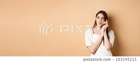 Dreamy and romantic girl looking aside with thoughtful gaze, touching clean gentle facial skin, standing against beige background Dreamy and romantic girl looking aside with thoughtful gaze, touching clean gentle facial skin, standing against beige background 103145211