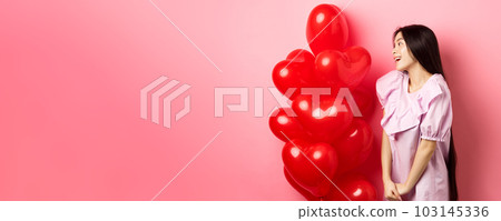 Romance and love concept. Romantic asian girl looking with affection and sympathy at empty space, standing near red hearts balloons from lover, pink background 103145336