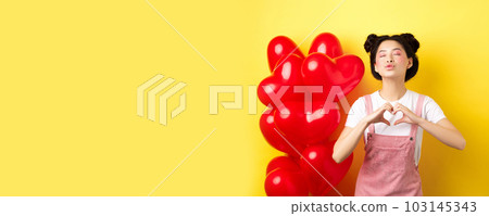 Romantic girl close eyes and pucker lips for kiss, showing I love you heart gesture, standing near cute red balloons, kissing lover over yellow background Romantic girl close eyes and pucker lips for kiss, showing I love you heart gesture, standing near cute red balloons, kissing lover over yellow background 103145343