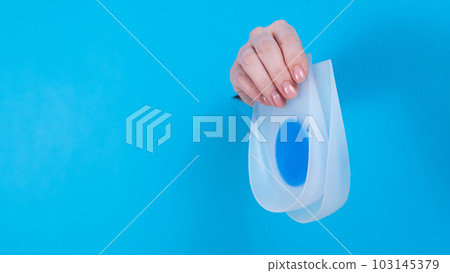 Woman holding orthopedic silicone heel pads. A woman's hand sticks out of a hole in a cardboard blue background. 103145379