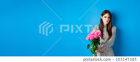 Valentines day concept. Displeased young woman rejecting flowers gift, frowning and grimacing, refusing take roses, standing on blue background Valentines day concept. Displeased young woman rejecting flowers gift, frowning and grimacing, refusing take roses, standing on blue background 103145385