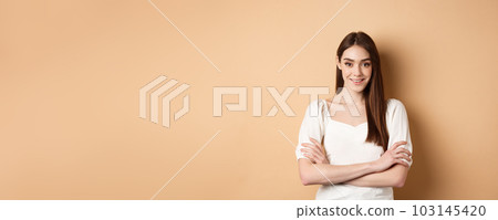 Smiling candid girl in dress cross arms on chest, looking like professional, standing confident on beige background Smiling candid girl in dress cross arms on chest, looking like professional, standing confident on beige background 103145420