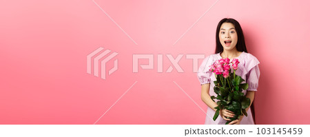 Korean teen girl in dress having romantic date on Valentines day, holding bouquet of roses and looking surprised at camera, receive gift on date from lover, pink background Korean teen girl in dress having romantic date on Valentines day, holding bouquet of roses and looking surprised at camera, receive gift on date from lover, pink background 103145459