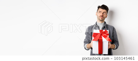 Pensive boyfriend looking aside and holding gift box, waiting for lover, making surprise present on valentines day, planning date with girlfriend, white background 103145461