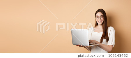 Smiling girl student working on laptop, wearing glasses and looking happy, using computer while standing against beige background 103145462