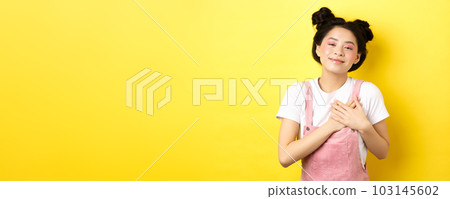 Beautiful teen asian girl in love, holding hands on heart and smiling touched, looking with affection, standing on yellow background Beautiful teen asian girl in love, holding hands on heart and smiling touched, looking with affection, standing on yellow background 103145602