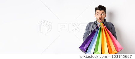 Excited man holding colorful shopping bags and looking left at special promo offer in store, standing on white background 103145697