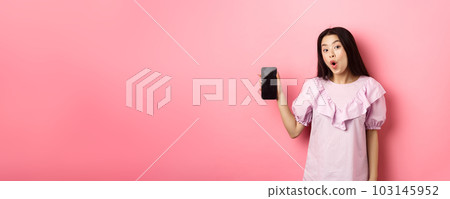 Online shopping. Excited asian woman say wow, showing empty smartphone screen and looking amazed at camera, standing against pink background Online shopping. Excited asian woman say wow, showing empty smartphone screen and looking amazed at camera, standing against pink background 103145952
