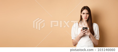 Disappointed girl with smartphone frowning, pucker lips upset, reading bad news on phone, standing on beige background 103146660