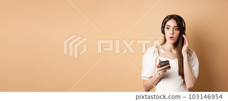 Wow awesome tunes. Excited young girl amazed with new song, listening music in headphones and look impressed, holding cell phone, beige background Wow awesome tunes. Excited young girl amazed with new song, listening music in headphones and look impressed, holding cell phone, beige background 103146954