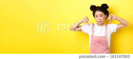 Asian beauty girl cringe from something disgusting, look away and showing thumbs-down to express dislike, negative emotions, standing in summer clothes on yellow background 103147003