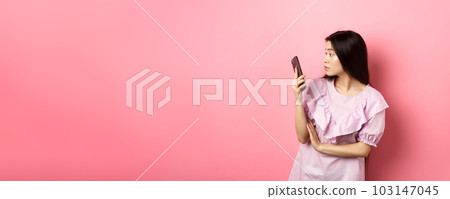 Asian girl look surprised at smartphone screen, reading message, standing in dress against pink background Asian girl look surprised at smartphone screen, reading message, standing in dress against pink background 103147045
