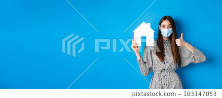 Real estate, covid-19 and pandemic concept. Cheerful woman in medical mask showing paper house cutout and thumbs up, recommend agency or realtor website, blue background Real estate, covid-19 and pandemic concept. Cheerful woman in medical mask showing paper house cutout and thumbs up, recommend agency or realtor website, blue background 103147053