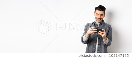 Handsome young man playing mobile video game and smiling at screen, standing against white background 103147125