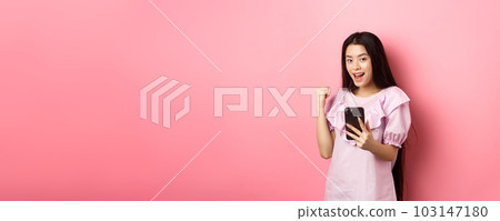 Online shopping. Satisfied asian girl winning on mobile phone, say yes and make fist pump, holding smartphone, standing against pink background 103147180