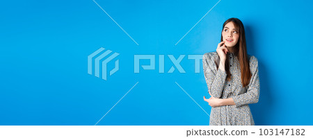 Creative young woman imaging and smiling, looking at upper left logo thoughtful, having an idea, standing on blue background in dress Creative young woman imaging and smiling, looking at upper left logo thoughtful, having an idea, standing on blue background in dress 103147182