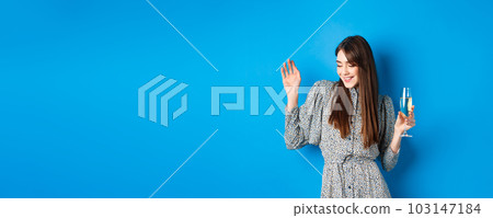 Celebration and party concept. Dancing girl holding glass of champagne, having fun at holiday, celebrating birthday, standing on blue background Celebration and party concept. Dancing girl holding glass of champagne, having fun at holiday, celebrating birthday, standing on blue background 103147184